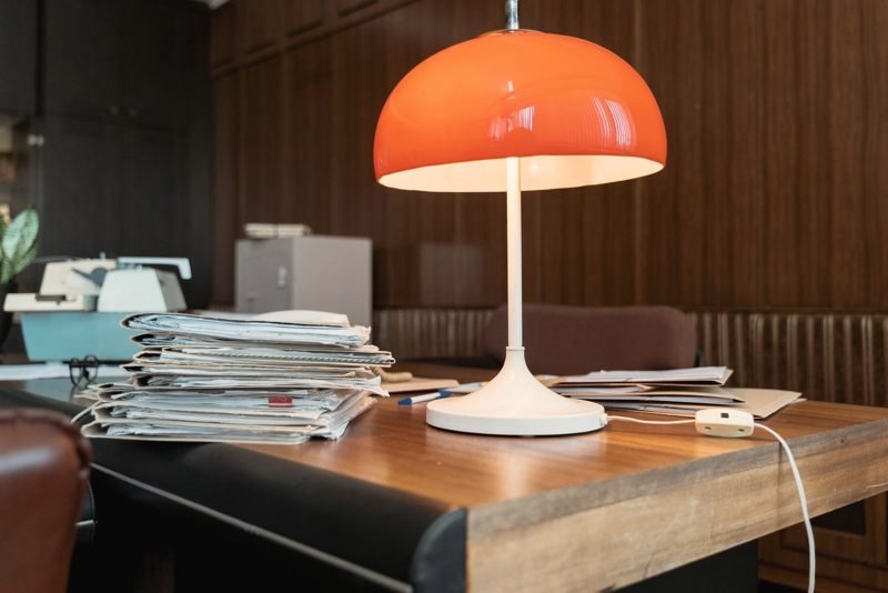 Stacks of paper files beside an orange desk lamp on a wooden office desk